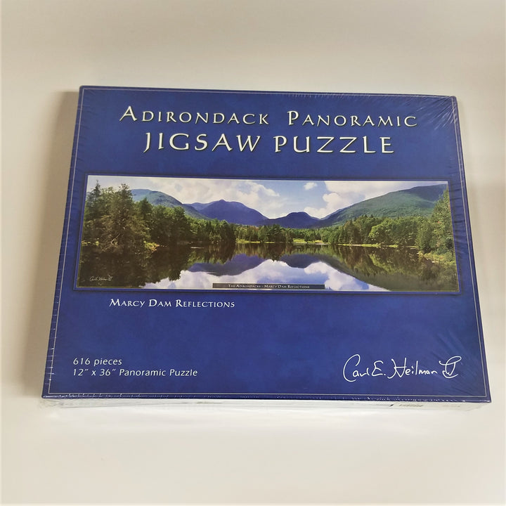 Box cover of Adirondack Panoramic Jigsaw Puzzle: Marcy Dam Reflections. Features a blue border around a rectangular photo of water surrounded by greenery with mountains in the background and reflected in the still water foreground.
