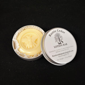 Open tin of Rustic Cedar Lotion Bar. To the left is the pale yellow circular bar with a bee sculpted in the center set in white crinkle paper. The lid of the tin is to the right. The top features the circular label with the Rustic Cedar logo in the center--two goats on either side of a tree. The ingredients are listed below that, the email of the crafter and the weight of the bar.