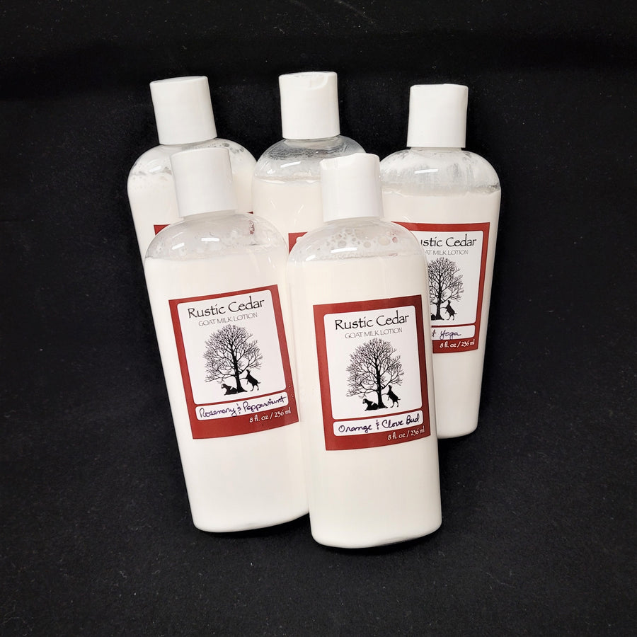 Five clear plastic bottles of various scents of Rustic Cedar Goat Milk Lotion grouped together on a black background. The lotion shows white through the bottle around the rust-bordered label in the center of the bottle. The label features the black logo in the center with two goats on either side of a tree. Over top is the text Rustic Cedar Goat Milk Lotion and below the logo the name of the scent. The two up front are Rosemary & Pepperment and Orange & Clove