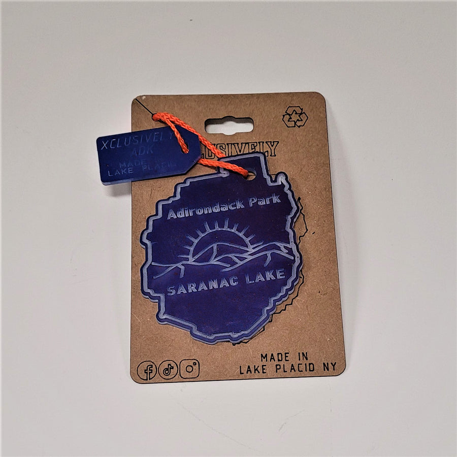 Single Adirondack Park ornament on its beige backing. The orange cord connects with a Made in Lake Placid tag to the dark blue ornament with Adirondack Park Lettering above a sunrise and mountains over the lettering SARANAC LAKE