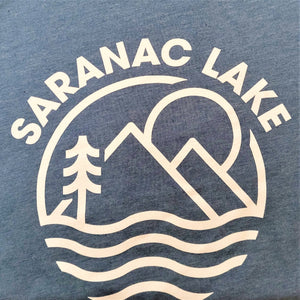 Close-up of white lettering and logo on the blue-gray t-shirt. SARANAC LAKE letters arc over a circular logo that includes a sun, mountains, evergreen and wavy lines for water.