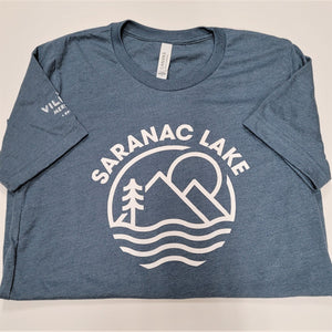 Folded gray-blue t-shirt. White lettering SARANAC LAKE arcs over a white logo of sun, mountains, evergreen and water. Short sleeves are turned in to reveal a vew white letters on the right sleeve. VIL MER