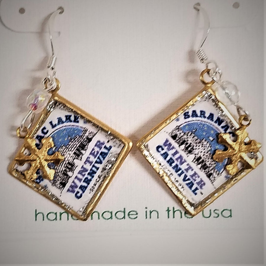 Dangling Winter Carnival square earrings. Gold band surrounds Saranac Lake Winter Carnival blue, white & black logo with small gold snowflake charm atop