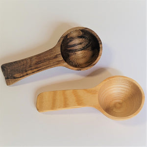Two wood coffee scoops on a white background. Top left is a dark grain scoop, below and slightly right is a light grain scoop.
