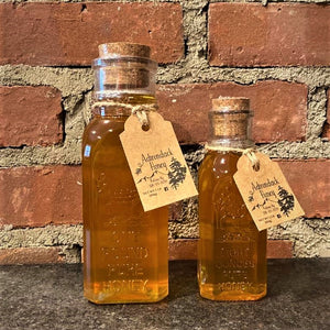 Two clear glass bottles of Adirondack Honey: one pound on the left; eight ounces on the right. Each bottle has a cork stopper and a tag with Adirondack honey logo depicted. Bottles are standing in front of a brick wall.