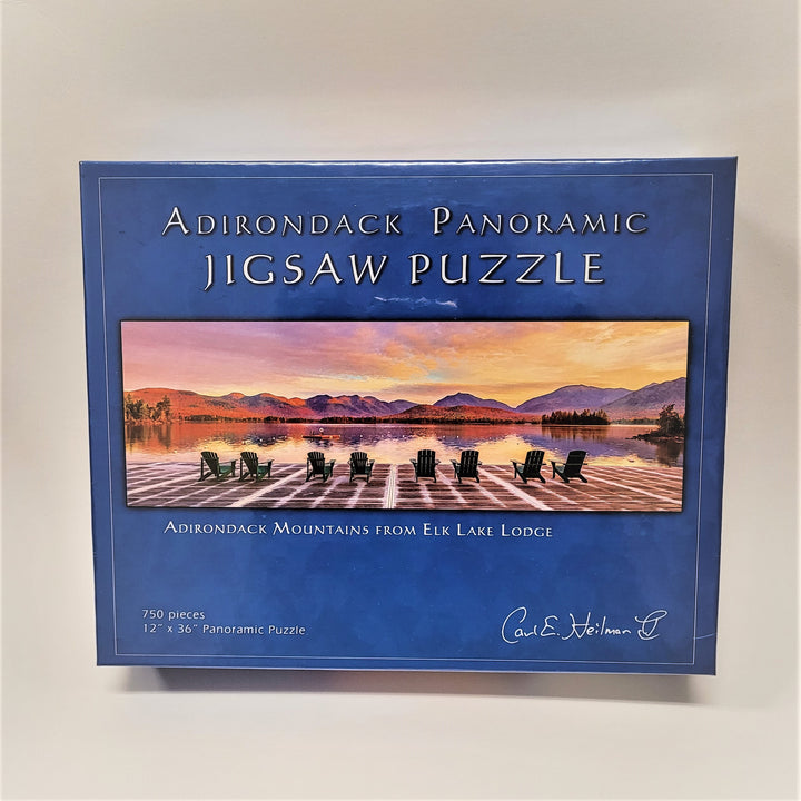 Puzzle box cover standing upright on a white background. Blue box with white type around the rectangular photo in the center of Adirondack chairs facing a slightly foggy pool of water with mountains in the background.