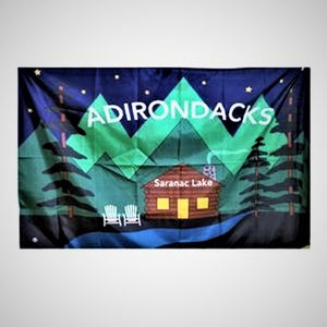 Flag opened to its full size with letters ADIRONDACKS in white type across green mountain tops and night sky over a log cabin with white text that reads Saranac Lake. The cabin's doors and windows are solid yellow. Two white Adirondack chairs sit to the left of the cabin . A ribbon of blue water is in front of the cabin with evergreens framing both sides of the flag.