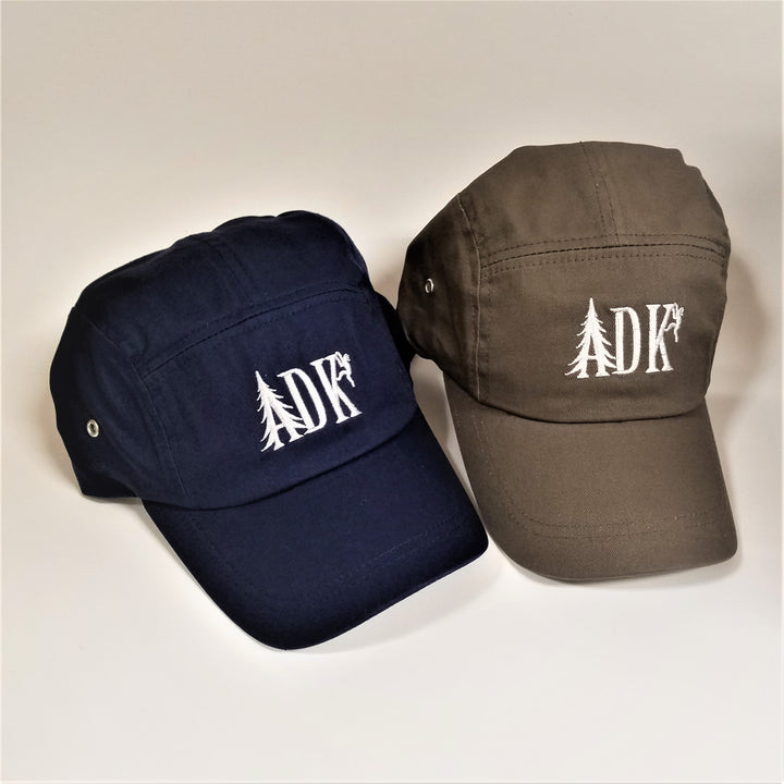 Two ADK panel caps. On the left a navy cap and to the right a brown cap. Both have white embroidery featuring  the letters ADK with the A in the style of a pine tree and a small hiker climbing the top of the K