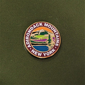 Close up of ADIRONDACK MOUNTAINS circular pin with colorful center circle depicting mountain, water and greenery. Outer circle has white type ADIRONDACK MOUNTAINS * NEW YORK. Pin is centered on the green card it is attached to.