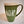 Tall green mug with whie stripe around the top with white imprint featuring the letters ADK with the A in the style of a pine tree and a small hiker climbing the top of the K.  Extra large handle visible on the right side.