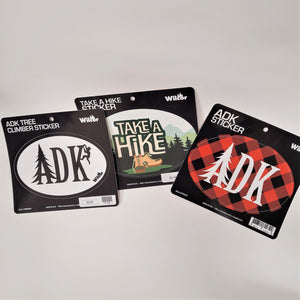 Three stickers fanned out on their packaging cards. Each is oval. Furthest left is black and white with ADK in black lettering with the A looking like an evergreen and a climber on the K. Middle sticker says TAKE A HIKE in white with green outline surrounded by mountains and evergreens and beige hiking boots below the lettering on top of a green swath. Furthest right is a buffalo plaid oval sticker with ADK in white type with the A looking like an evergreen.