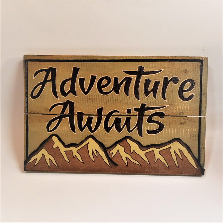Front view of wooden sign with Adventure Awaits type in black on a board with a horizontal alit in the word Awaits over snow-covered mountains across the bottom of the sign