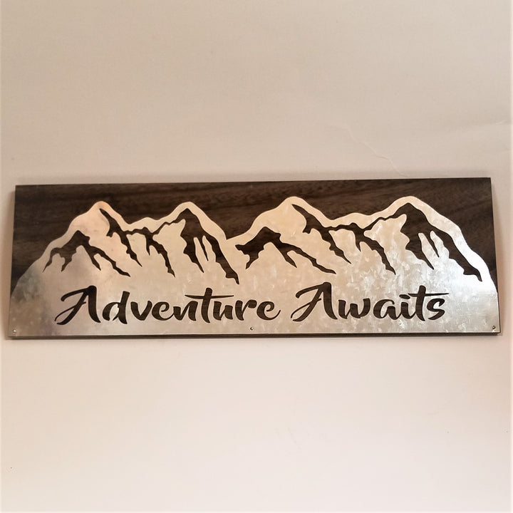 Rectangular sign with dark brown wood backing and silver plate mountains with brown showing through the peaks and the word Adventure Awaits cut through.