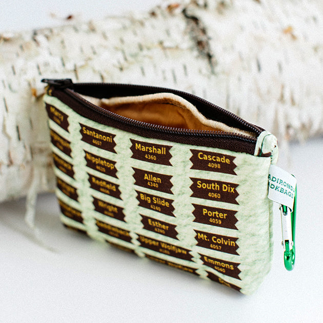 Zipper change purse leaning on birch log. Mountain names and elevations are printed on brown marker-like signs on the front of the bag.