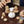 Fingers carefully holding tiny wooden loaf of bread aiming for the bakers pile on the left. The white-wooden rounded baker is centered on a wooden table with various tiny wooden baked goods surrounding him and another set of fingers holding a tiny wooden pie coming from the right.