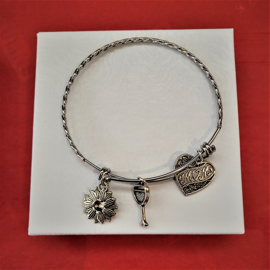 Single bangle bracelet: silver ring with three charms hanging below: flower with petals, wine glass, and heart with MOM in a center banner. All set n a white box lid on a red background.