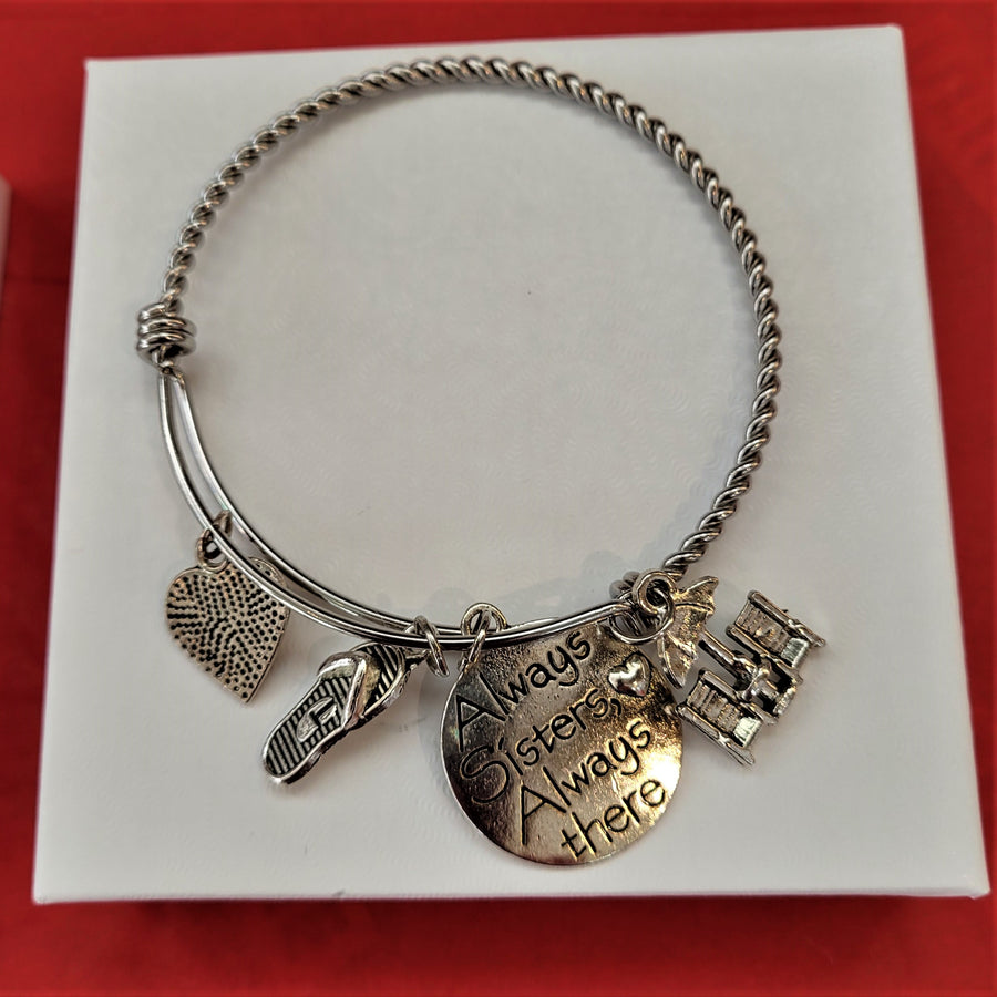 Single bangle bracelet: silver ring with four charms hanging below: a heart with lots of dots, a flip flop, a sphere that says Always Sisters Always there and a beach umbrella with two beach chairs below. All set n a white box lid on a red background.