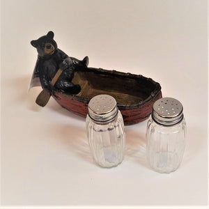 Bear with brown paddle on left side of red canoe with clear glass salt and pepper shakers outside of canoe in the front and on the right.