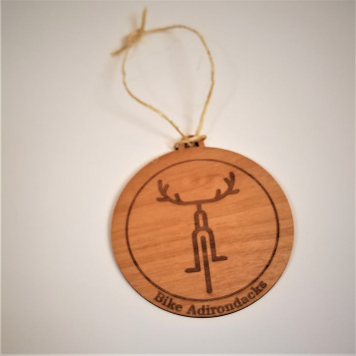 Faux wood round ornament with brown bicycle icon with antler-like handlebars within a brown circle. Below the circle in brown type are the words Bike Adirondacks.  A thin-rough cord is attached to the top for hanging.