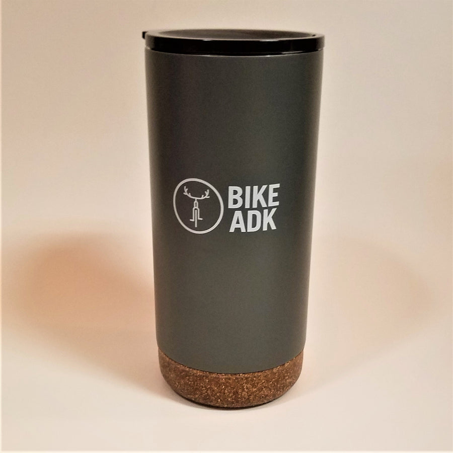 Charcoal tumbler with cork-like bottom stands upright with the text BIKE ADK in white lettering and the iconic bicycle with antler handlebars in a white circle next to the type. A thin black lid is visible at the top of the tumbler.