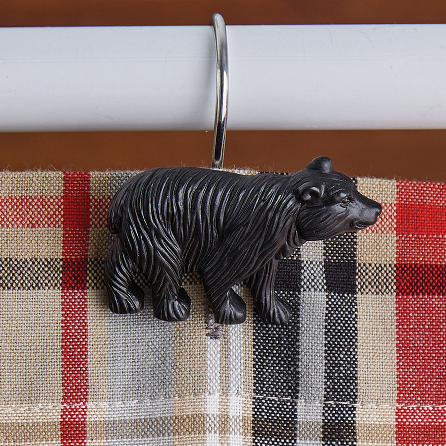 Black bear sits on the end of a metal shower hook which is hung over a white pole. Beneath the black bear is a red, black, tan and white plaid curtain.