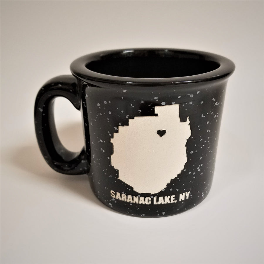 A single black-with-gray speckled camp mug with the words SARANAC LAKE, NY on the bottom and a white-filled outline of the Adirondack Park in the center. There is a small black heart in the white outline located where Saranac Lake sits. The mug has a black rim and black interior.