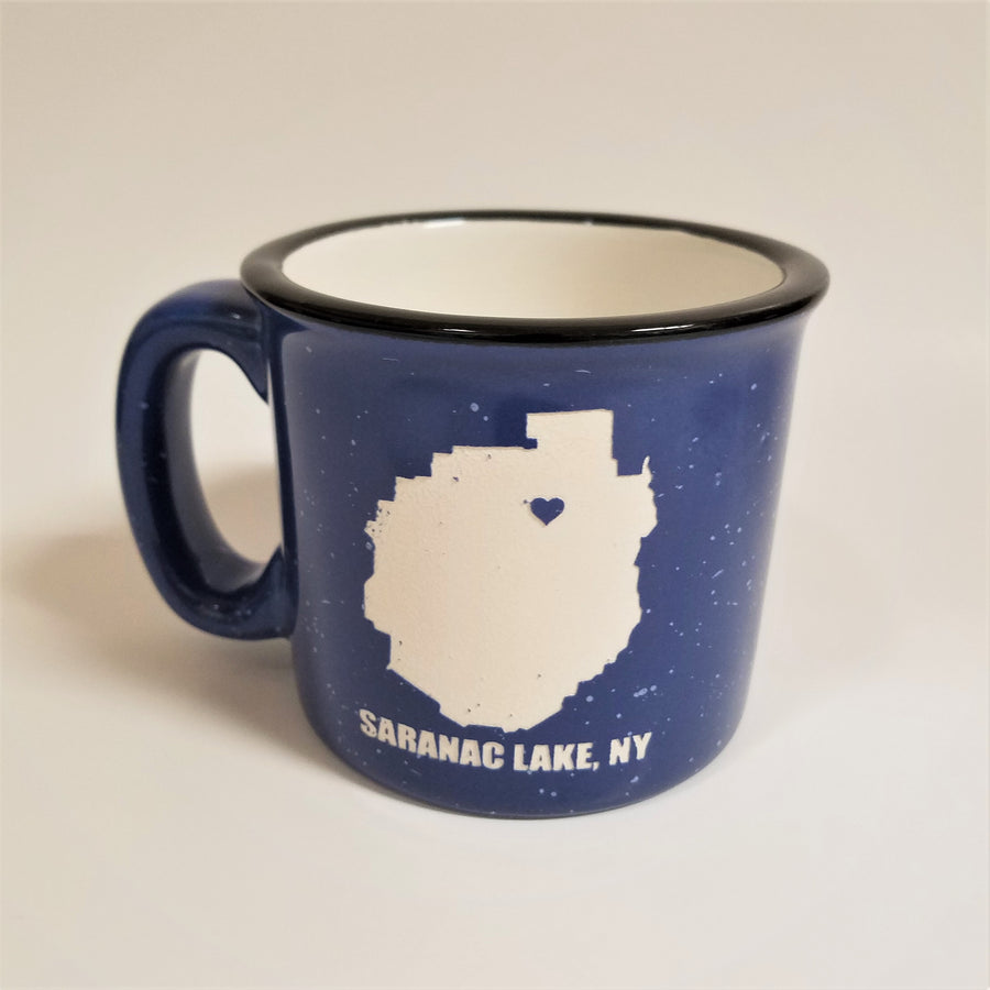 A single blue-with-white speckled camp mug with the words SARANAC LAKE, NY on the bottom and a white-filled outline of the Adirondack Park in the center. There is a small blue heart in the white outline located where Saranac Lake sits. The mug has a black rim and white interior.