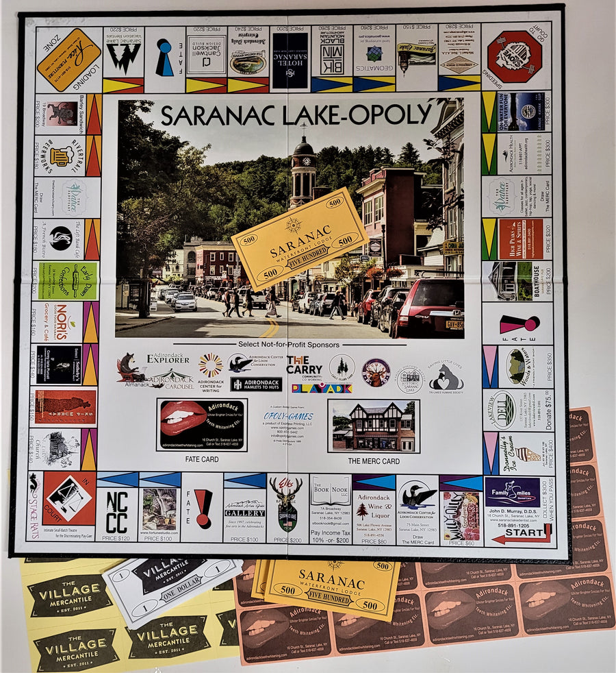 Game board of SARANAC LAKE-OPOLY open with game money on the board and below along with Merc Cards and Fate Cards featured below the board.
