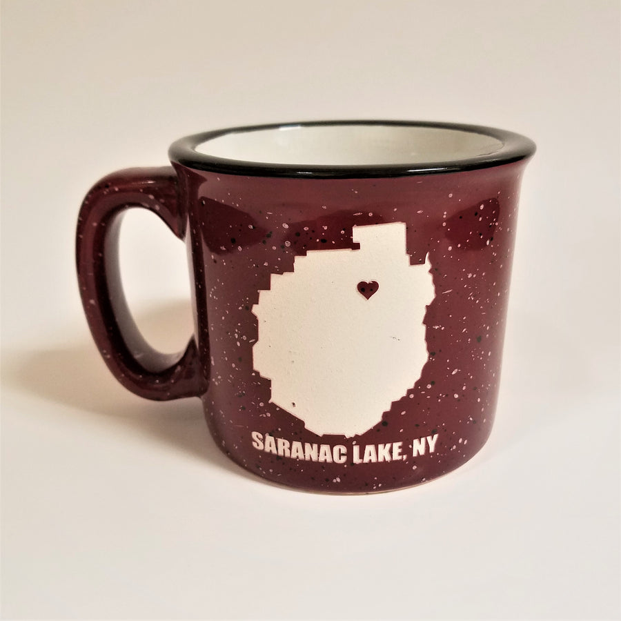 A single burgundy-with-white speckled camp mug with the words SARANAC LAKE, NY on the bottom and a white-filled outline of the Adirondack Park in the center. There is a small burgundy heart in the white outline located where Saranac Lake sits. The mug has a black rim and white interior.