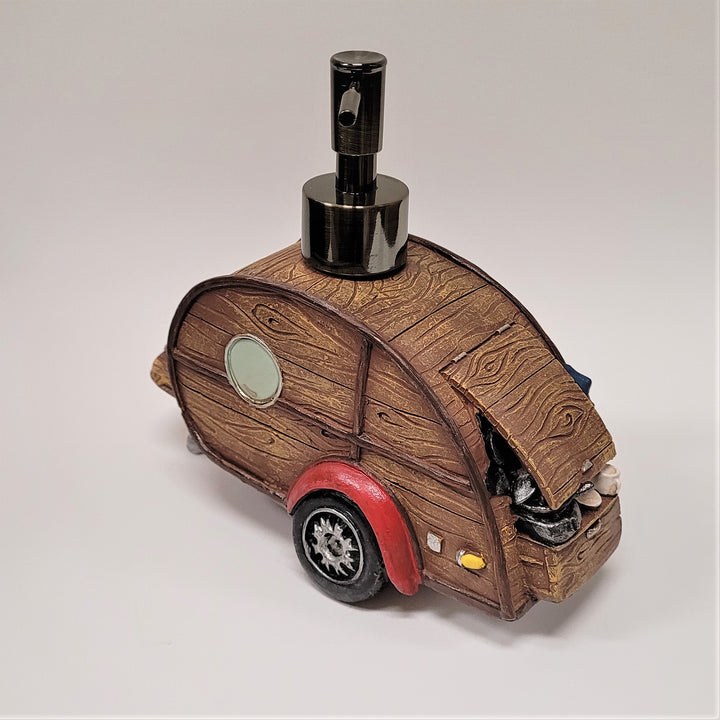 Woody camper soap dispenser slightly angled on a white background.  The metal push spout sits atop the brown wood-grained look o fthe camper complete with hatch, circular window and red shield with wheel beneath.