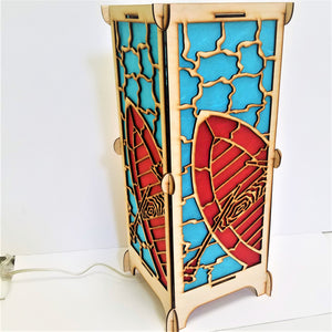 Art lamp standing straight up revealing two panels of the four-sided lamp. Red boat and oars, blue water and light wood creating the outlines and overall structure. Cord comes from bottom and extends to the left.