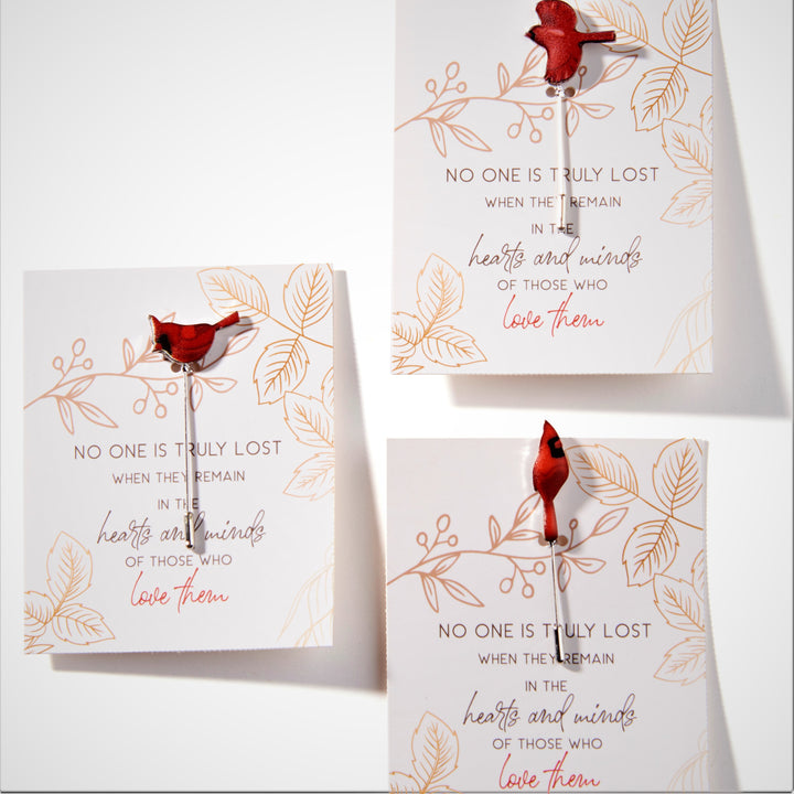 3 different cardinal stick pins on the sentiment card they are sold with. Cardinal on left is in profile with wings closed. Bottom right cardinal is upright front faced and cardinal on top has wings spread in flight. 