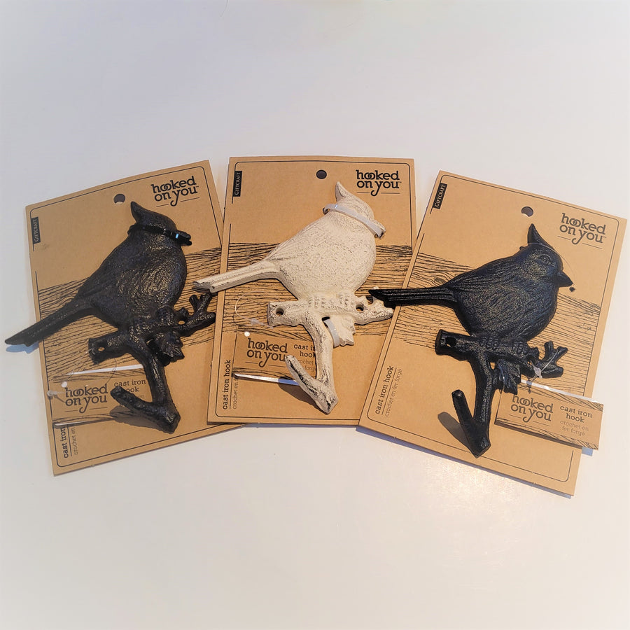 Three cast-iron cardinal wall hooks attached to their beige card packaging. On the left is the antique brown, middle antique white and right black birds on branch with hook below. All on a white background.