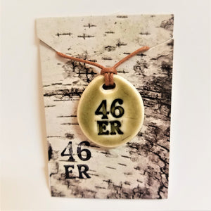 Ceramic, oval pendant with 46er in bold black in center and a taupe glaze around it on top of white. The ceramic pendant sits on a faux birch paper package with the brown necklace twine seen coming out of the birch card.