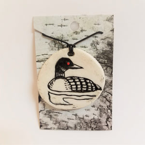 Circular white ceramic pendant with a red-eyed loon in the center. Set on faux-birch paper with the black necklace cord coming through.
