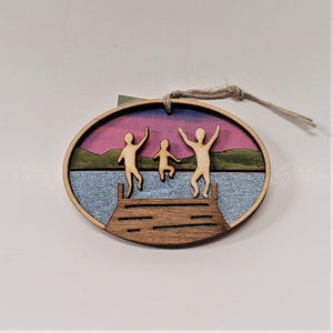 Three natural wooden children figures jumping off a brown wooden dock are featured in the center of this oval ornament. Silver blue water surrounds the dock with green mountains and purple sky in the background.