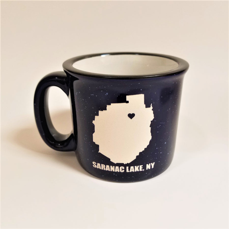 A single dark blue-with-white speckled camp mug with the words SARANAC LAKE, NY on the bottom and a white-filled outline of the Adirondack Park in the center. There is a small dark blue heart in the white outline located where Saranac Lake sits. The mug has a black rim and white interior.