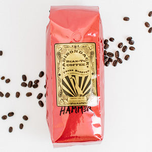 Red bag of Adirondack Bean-to-Bean Hammer coffee. The bag is standing on a white background with loose coffee beans strewn on either side.