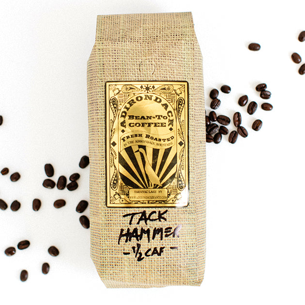 Faux-burlap bag of Adirondack Bean-to-Bean Tack Hammer 1/2 caf coffee. The bag is standing on a white background with loose coffee beans strewn on either side.
