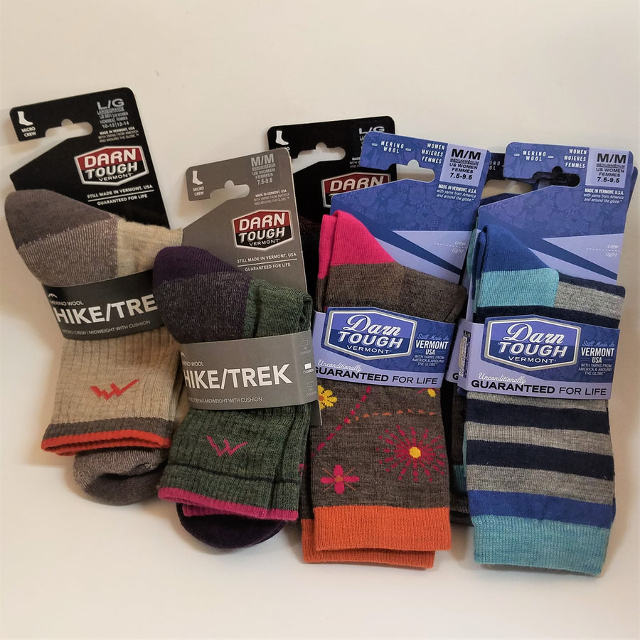 Full view of 4 pairs of Darn Tough socks in their packaging. Left to right: Hike/Trek sock in black labeled packaging--sock is pale beige with brown heel and brown and red stripes on the cuff; Hike/Trek  sock in black packaging--sock is dark gray with navy heel and pink and navy strip at the cuff; Blue labeled Darn Tough Women's solid brown with red, yellow, pink flowers and dotted lines, pink heel, orange cuff; Darn Tough Women's sock striped--blue, gray, black with a blue heel and aqua blue cuff