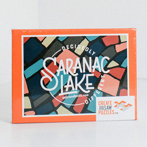 Box cover for Decidedly Different Saranac Lake jigsaw puzzle. Box border is rectangular orange around pieces in shades of blue, green, red, yellow with black borders.