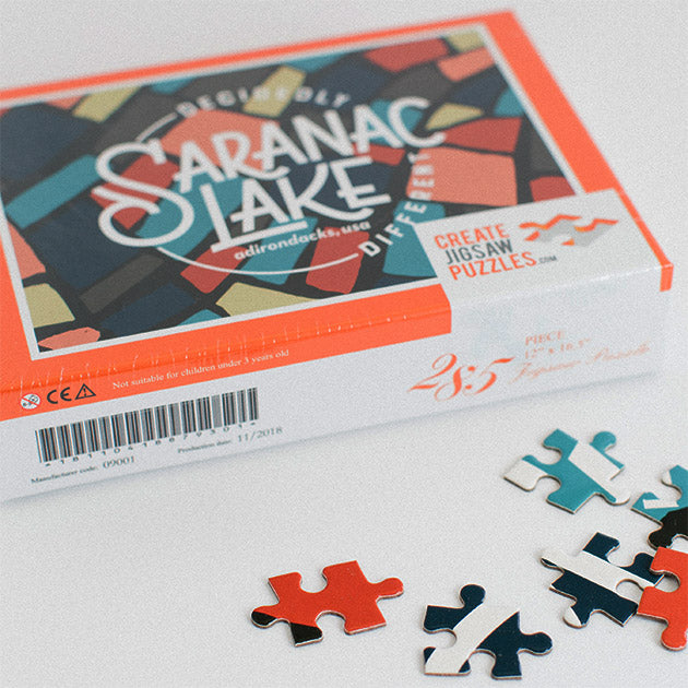 Decidedly Different Saranac Lake jigsaw puzzle box set further back with 5 loose pieces in front on a white background.