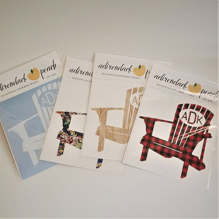 Fanned out display of Adirondack chair decals in white, floral,  wood grain, and buffalo plaid with an oval in top of chair with ADK lettering showing on some of the chair decals.