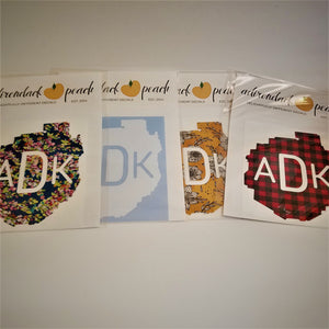 Fanned out display of Adirondack Park boundaries decals in navy floral pattern, white, yellow floral and buffalo plaid pattern with white ADK lettering printed in the middle of the decals