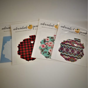 Fanned out display of Adirondack Park boundaries decals with white heart in top third slightly right center. From left to right partial white park outline on blue, buffalo plaid pattern, floral pattern and Aztec pattern.ern, white, yellow floral and buffalo plaid pattern .