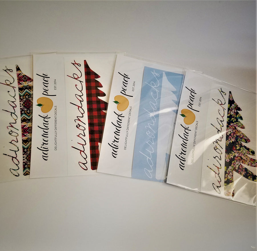 Fanned out display of Adirondack Peach half pine tree decals. Four packets display the pink aztec, buffalo plaid, white and navy floral patterns.