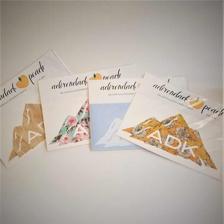 Fanned out display of Adirondack mountains decals in wood grain pattern, aqua floral, white,  and yellow floral with white ADK lettering printed at the base of the mountain range seen on some and peaking through.