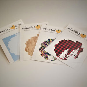 Fanned out display of Adirondack Park boundaries decals in white, wood grain, navy floral and buffalo plaid pattern with two white pine trees seen in full on the buffalo plaid pattern furthest to the right.