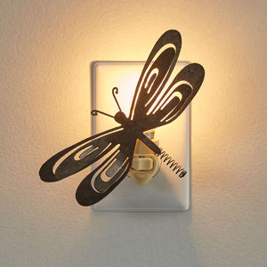 Metal dragonfly nightlight turned on in a plug with light emanating through its wings and above them.