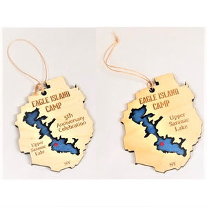 Two Eagle Island ornaments. with twine hanger above. Both are cream-colored with EAGLE ISLAND CAMP lettering on the top, and a blue lake cut out with a red dot for Eagle Island. Ornament on left says 5th Anniversary Celebration on the right with Upper Saranac Lake on the left. The ornament on the right says Upper Saranac Lake on the right. Both have the letters NY on the bottom.